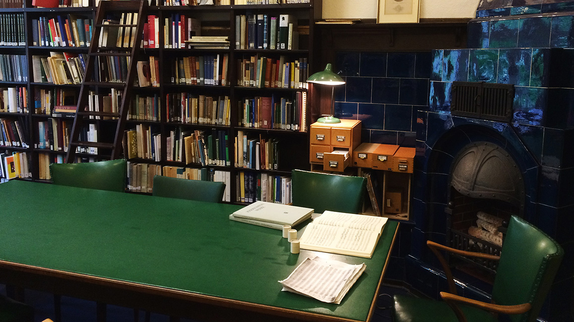 Blibliotheksregal mit Büchern, davor ein Tisch und Stuhl, auf dem Tisch liegen Papiere