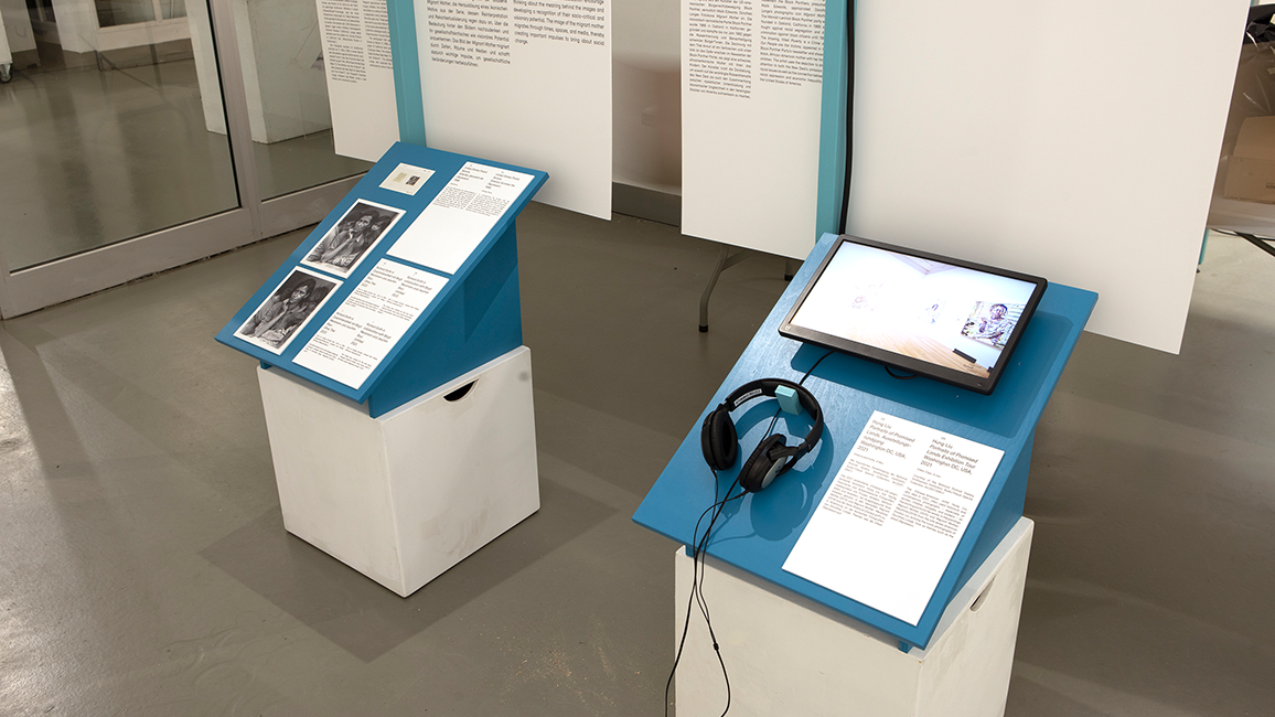 Ausstellungstische zu Migration. Kopfhörer für Audiobeschreibung liegt auf einem Tisch